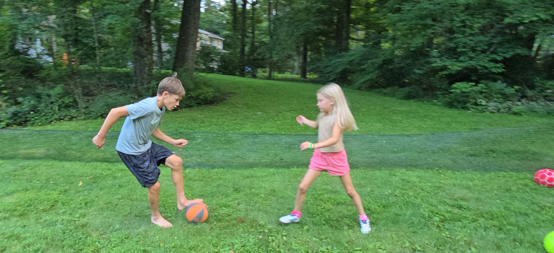 A boy shielding an orange ball from a girl closing him down, the perimeter net running behind them