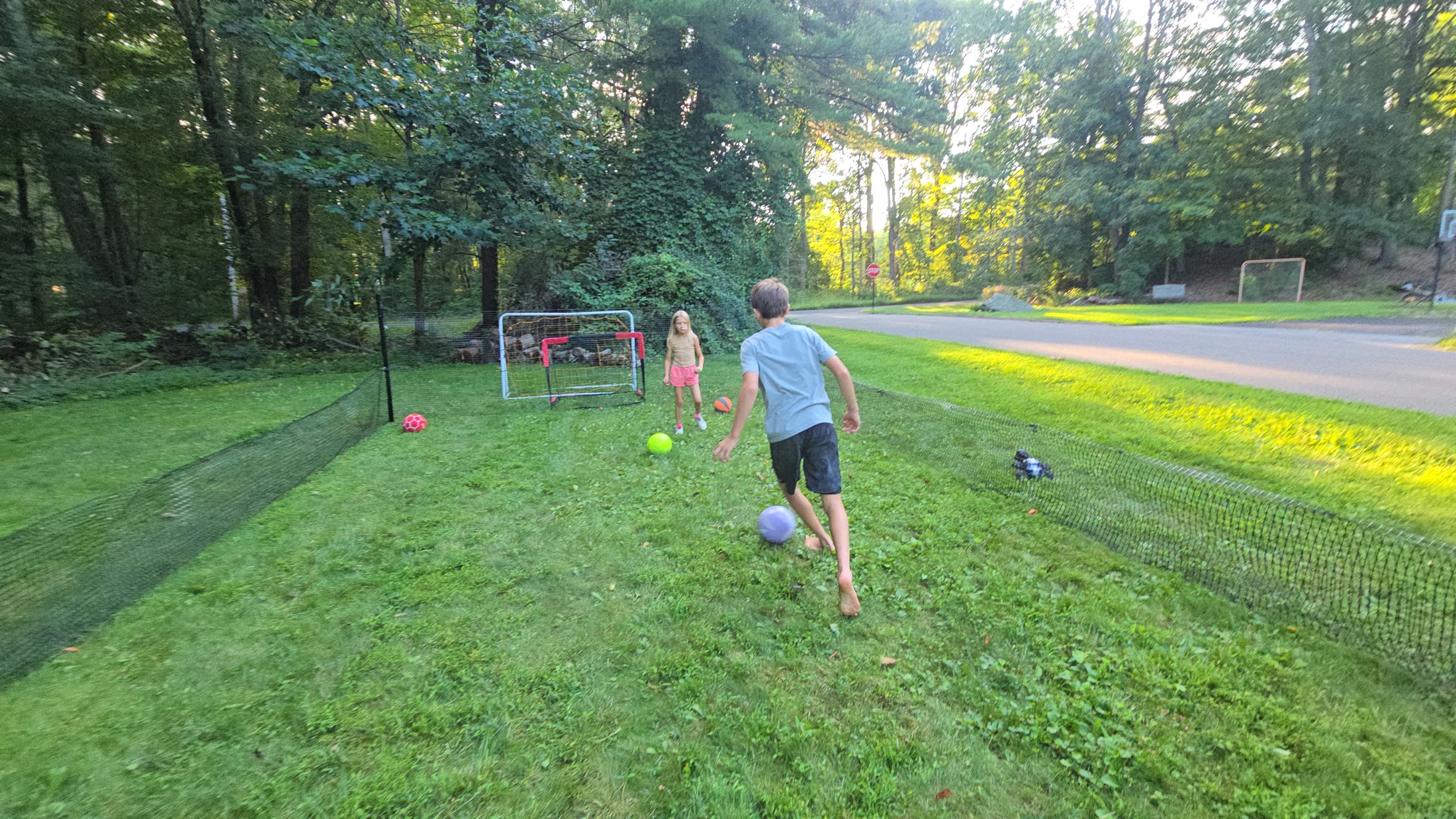 A boy running onto a ball inside a netted backyard field at golden hour while a girl waits by the goal