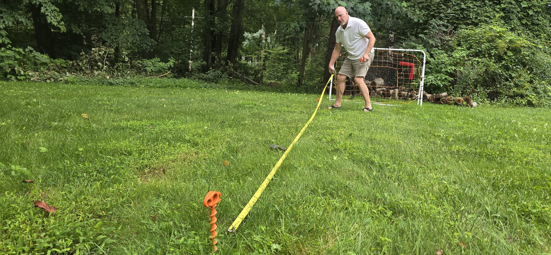 A man pulling a yellow tape measure across a lawn to set out the field, with a soccer goal in the background