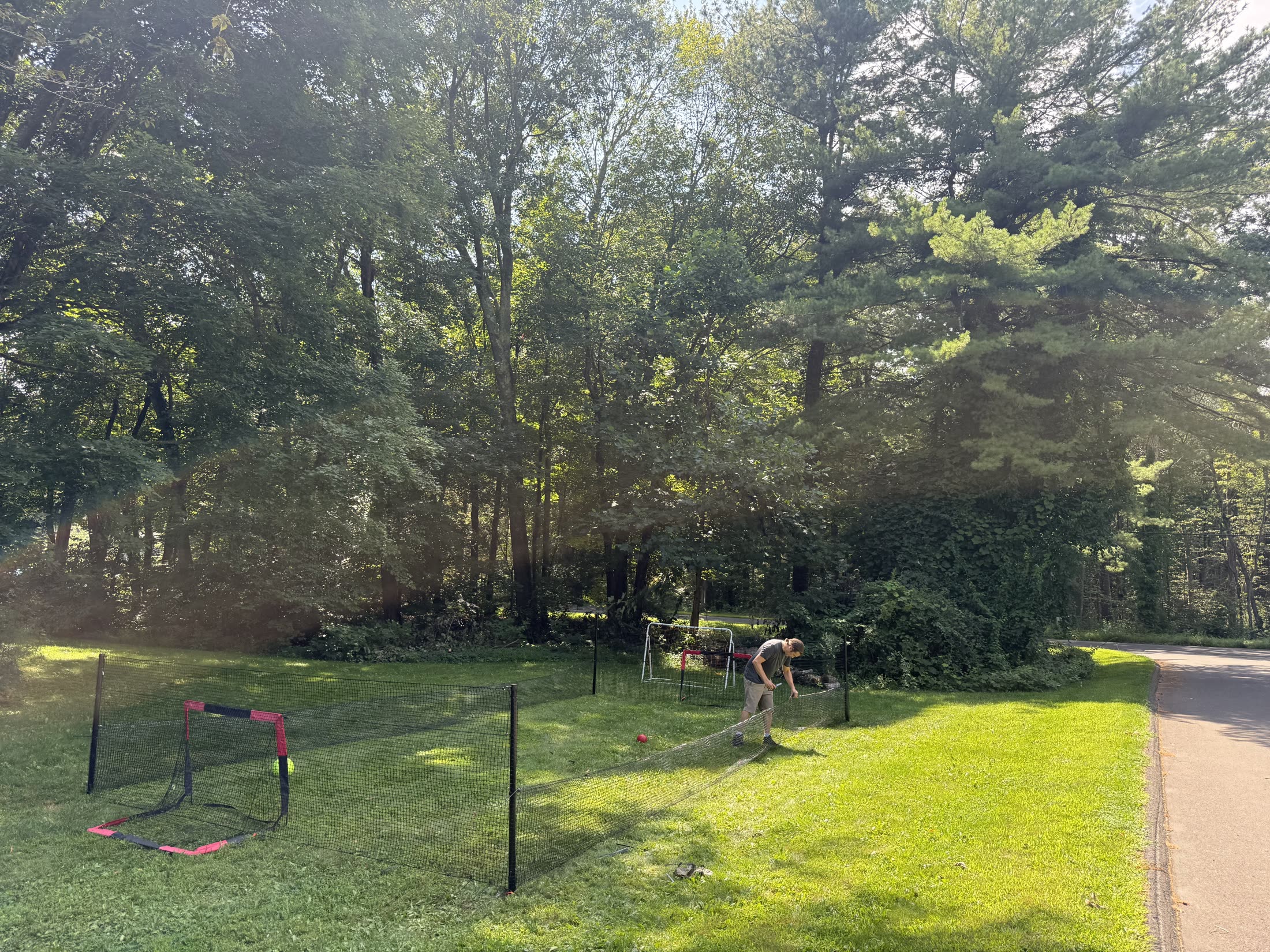 A man threading black perimeter netting onto a post along the edge of a sunlit lawn, with two small red-and-black goals already set up inside the enclosure