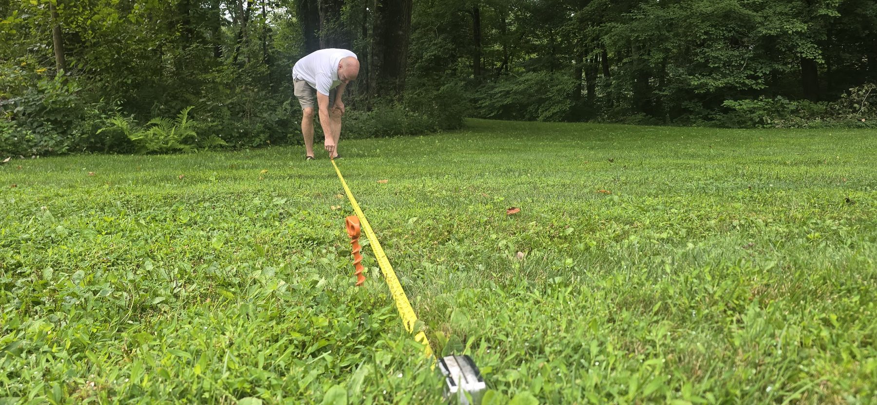 A man measuring across a wide lawn with a yellow tape and an orange anchor in the foreground