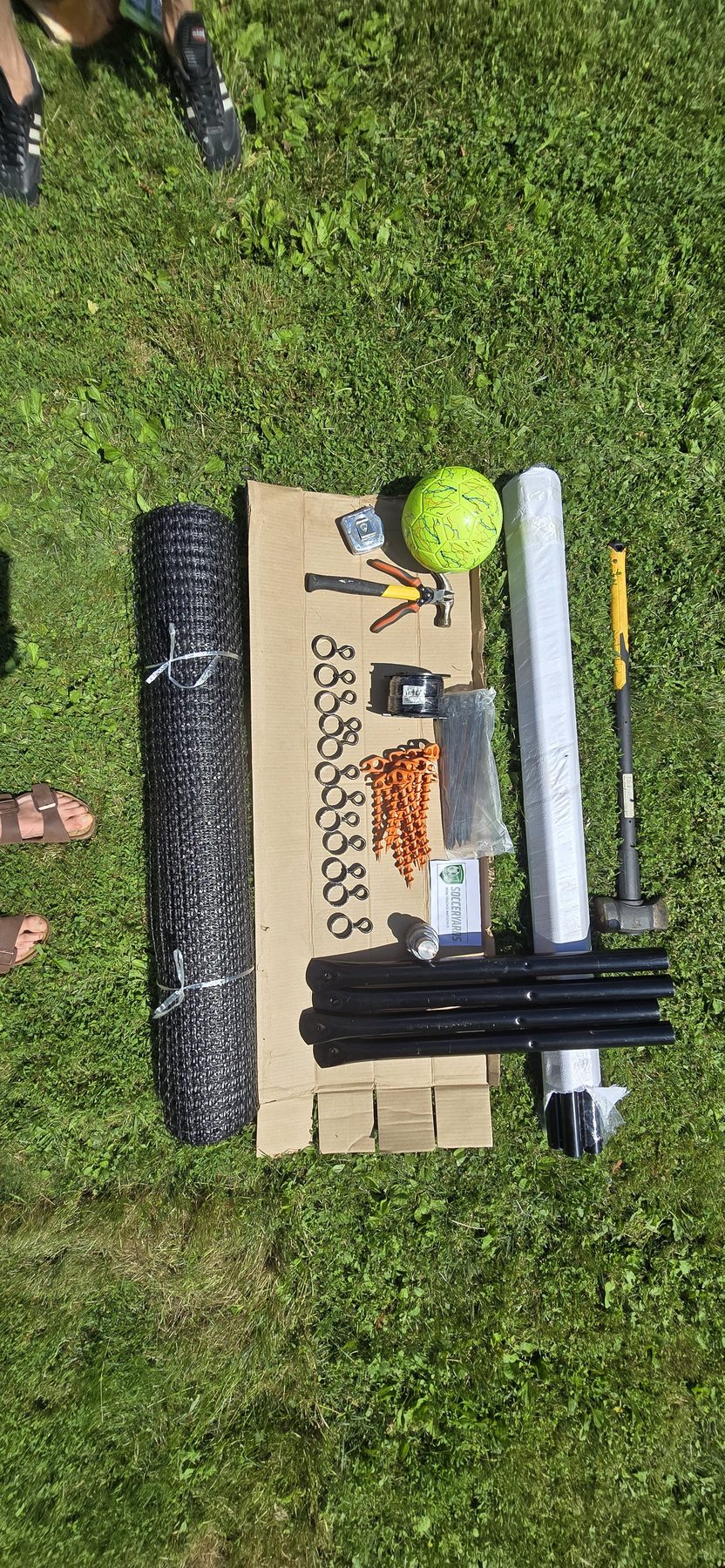 Overhead view of the SoccerYards kit on grass with the rolled wall panel, white and black posts and a yellow-green ball