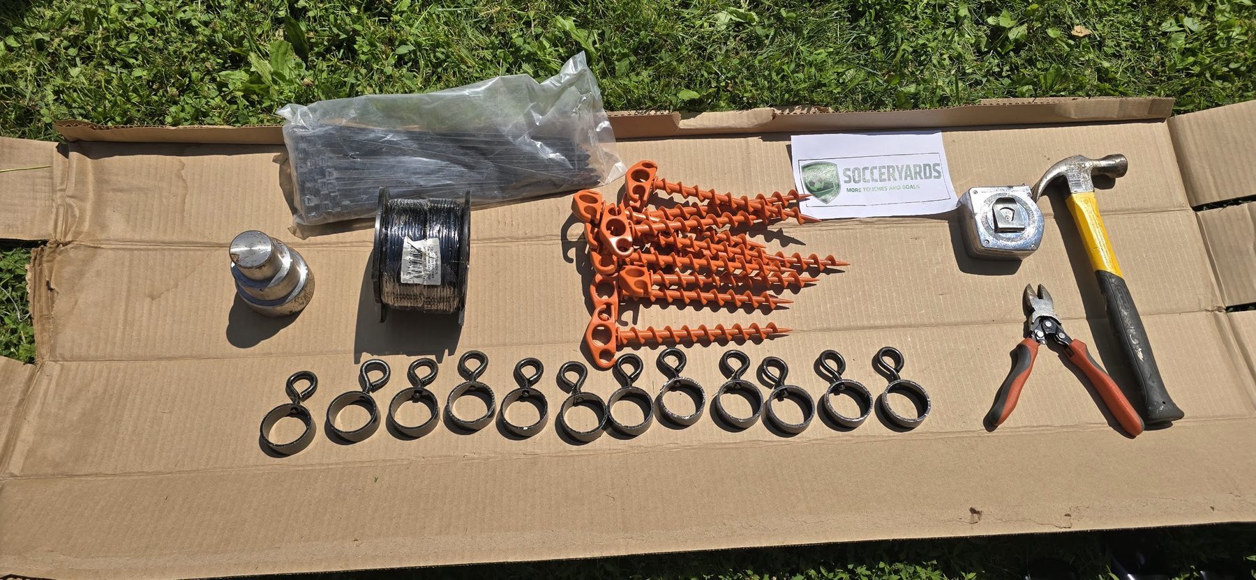 Close view of SoccerYards hardware arranged on cardboard: rubber washers, orange screw-in ground anchors, a tape measure and a hammer