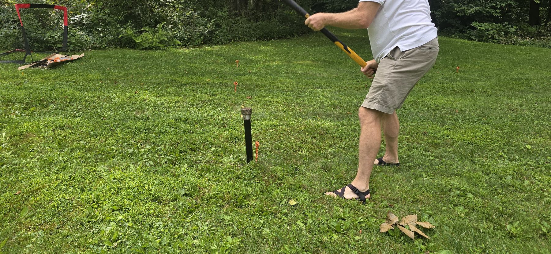 A man swinging a sledgehammer to drive a black ground sleeve into the lawn, orange markers running away behind him