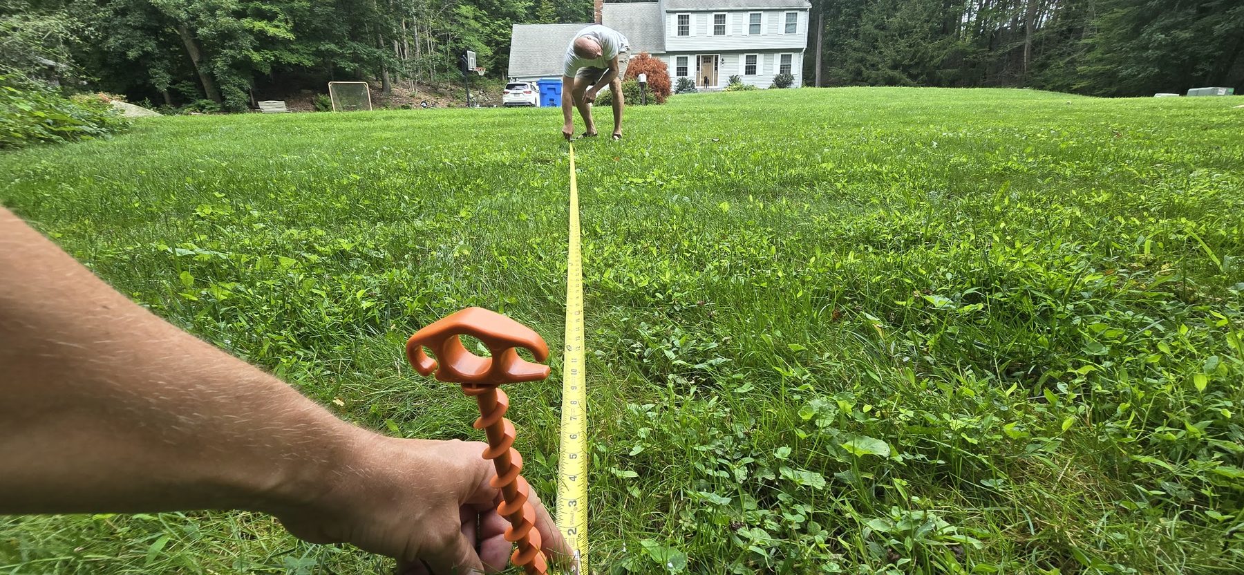 A hand holding an orange screw-in ground anchor next to a yellow tape measure stretched across the grass