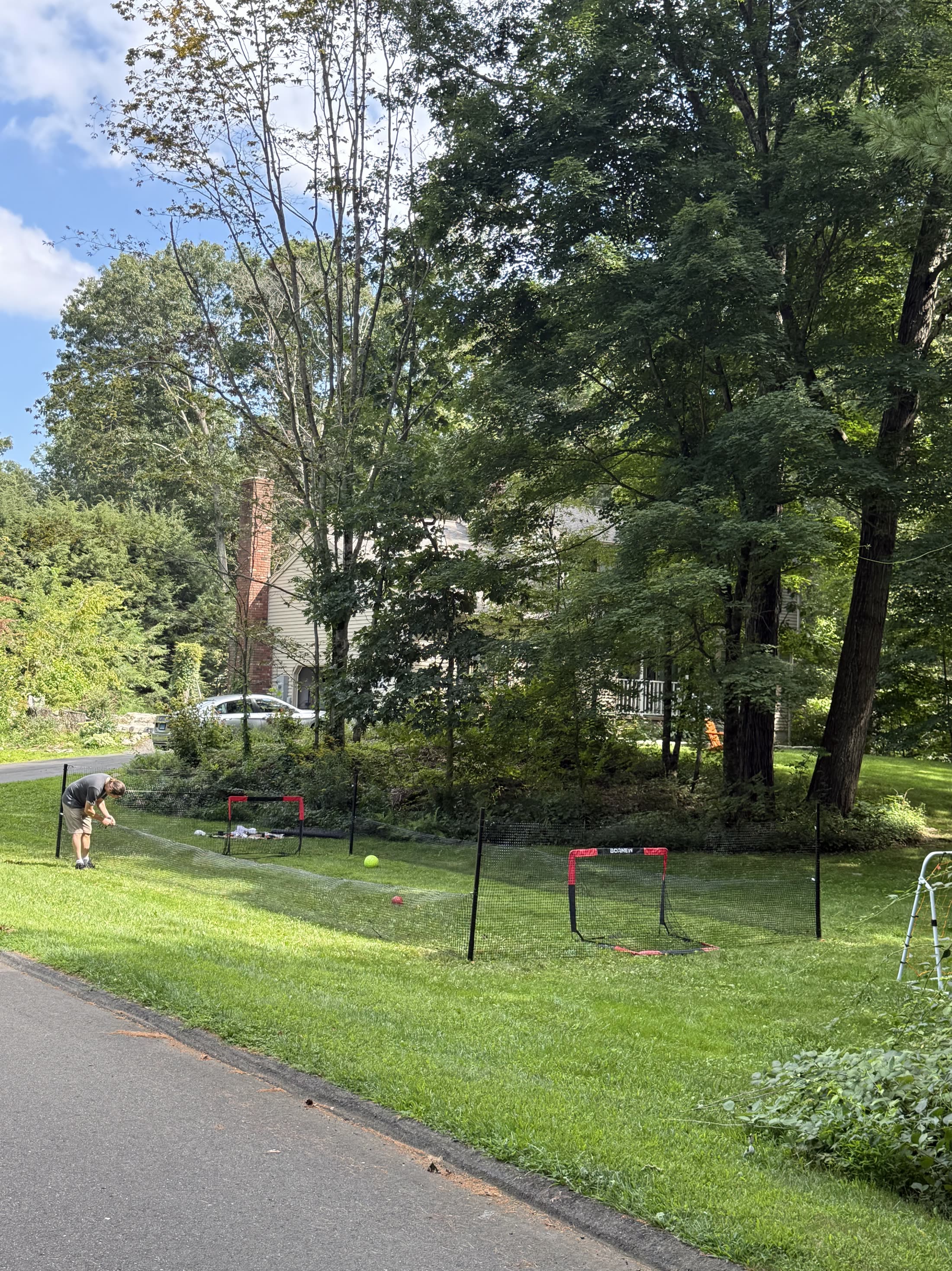 A man tensioning the perimeter net along a front lawn with two goals inside the enclosure and a house across the road