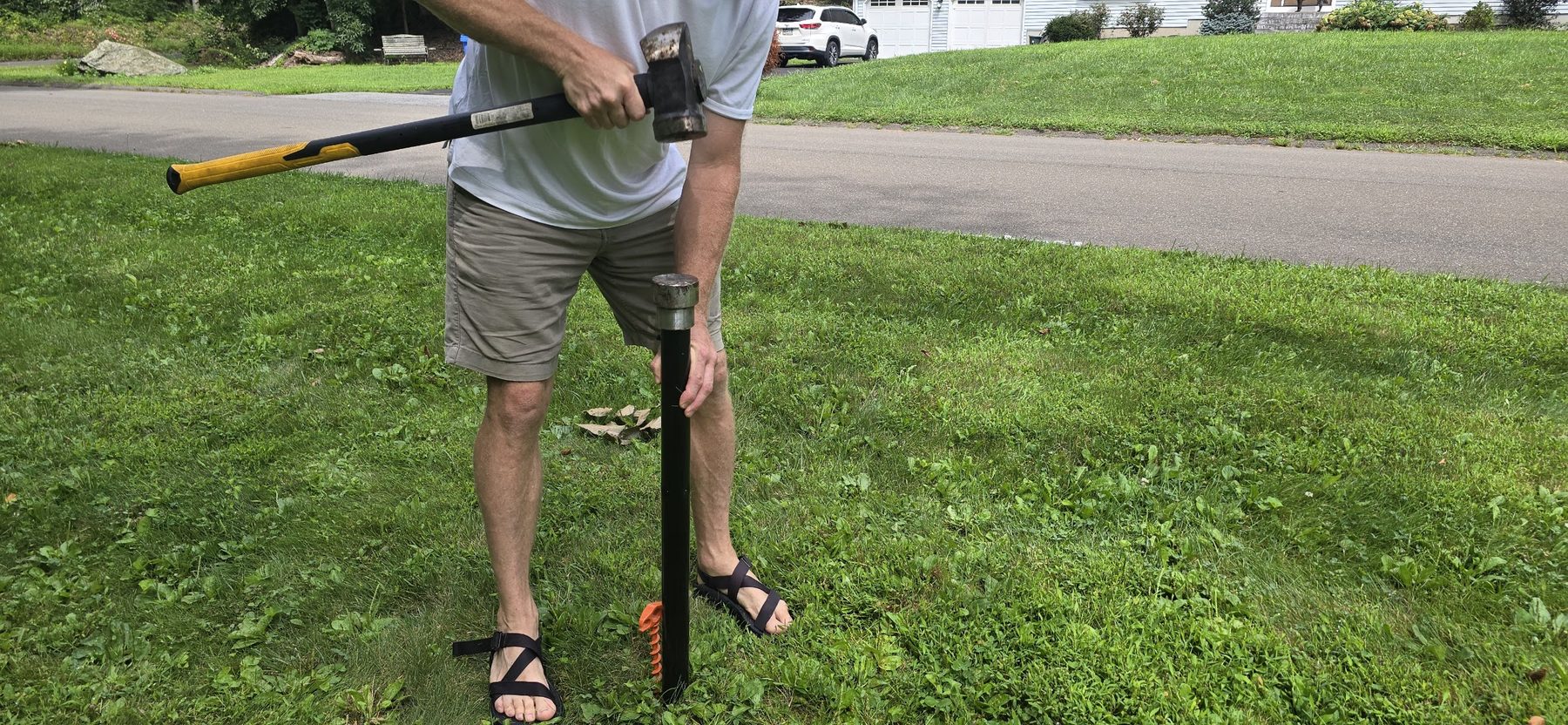 A man driving a ground sleeve into the lawn with a residential street visible behind him