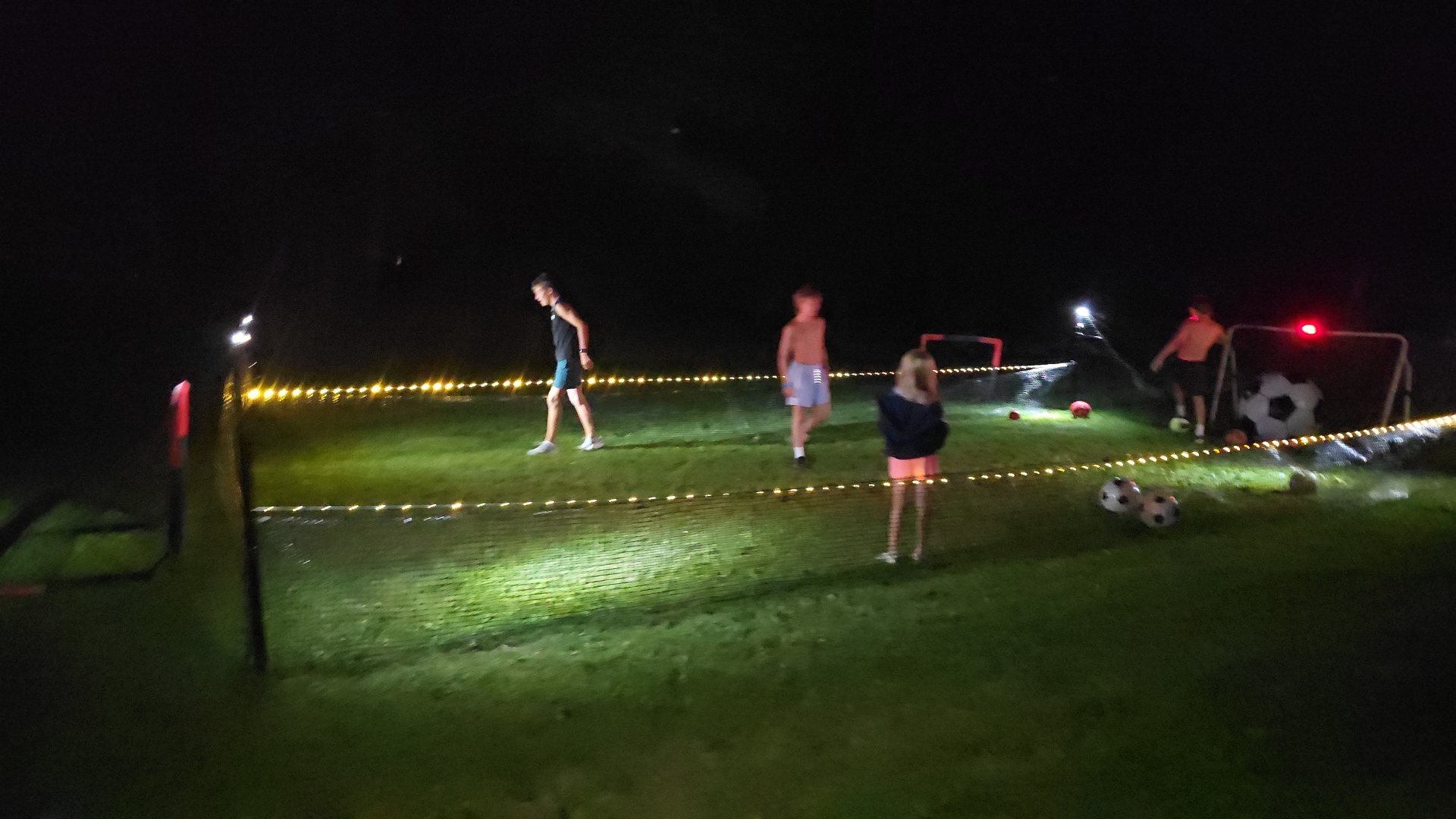Kids playing soccer at night inside a SoccerYards walled field lit by string lights