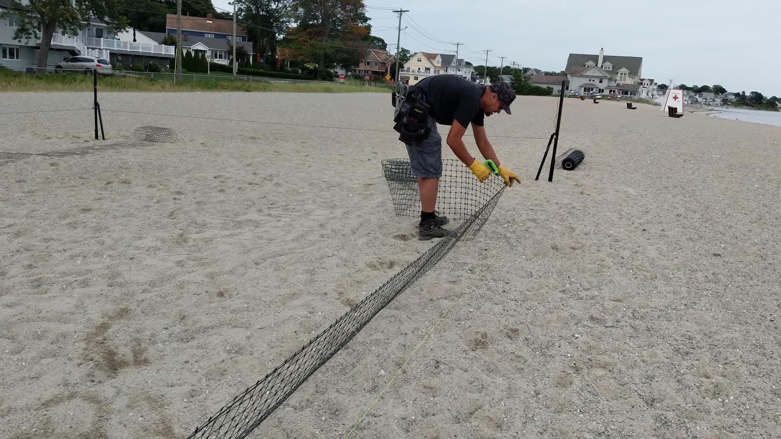 A man kneeling on the beach to fix netting to a post along a line of sand