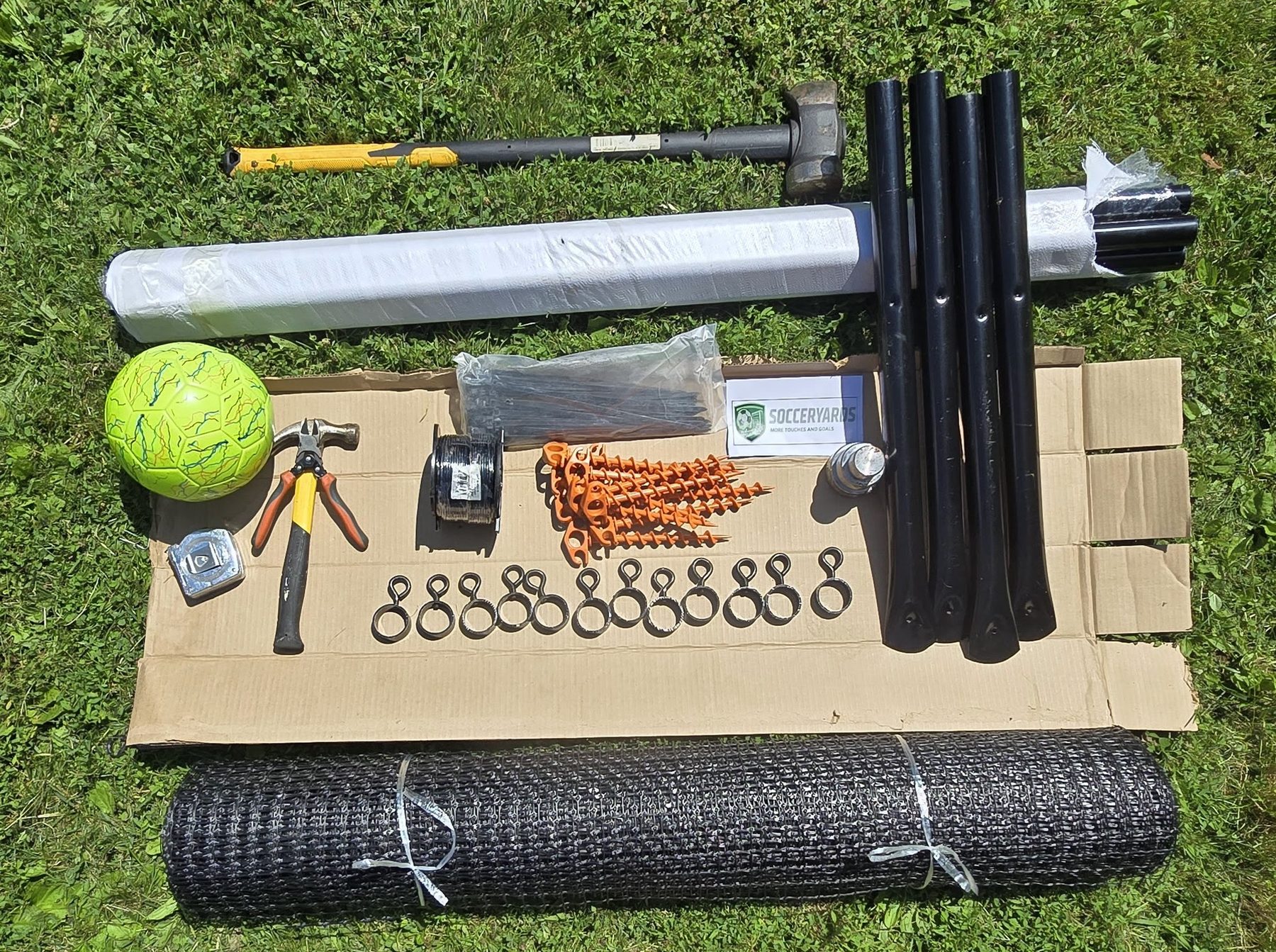 The full SoccerYards kit laid out on flattened cardboard in a mown backyard: black posts, a rolled wall panel, orange ground anchors, washers, a mallet and a ball