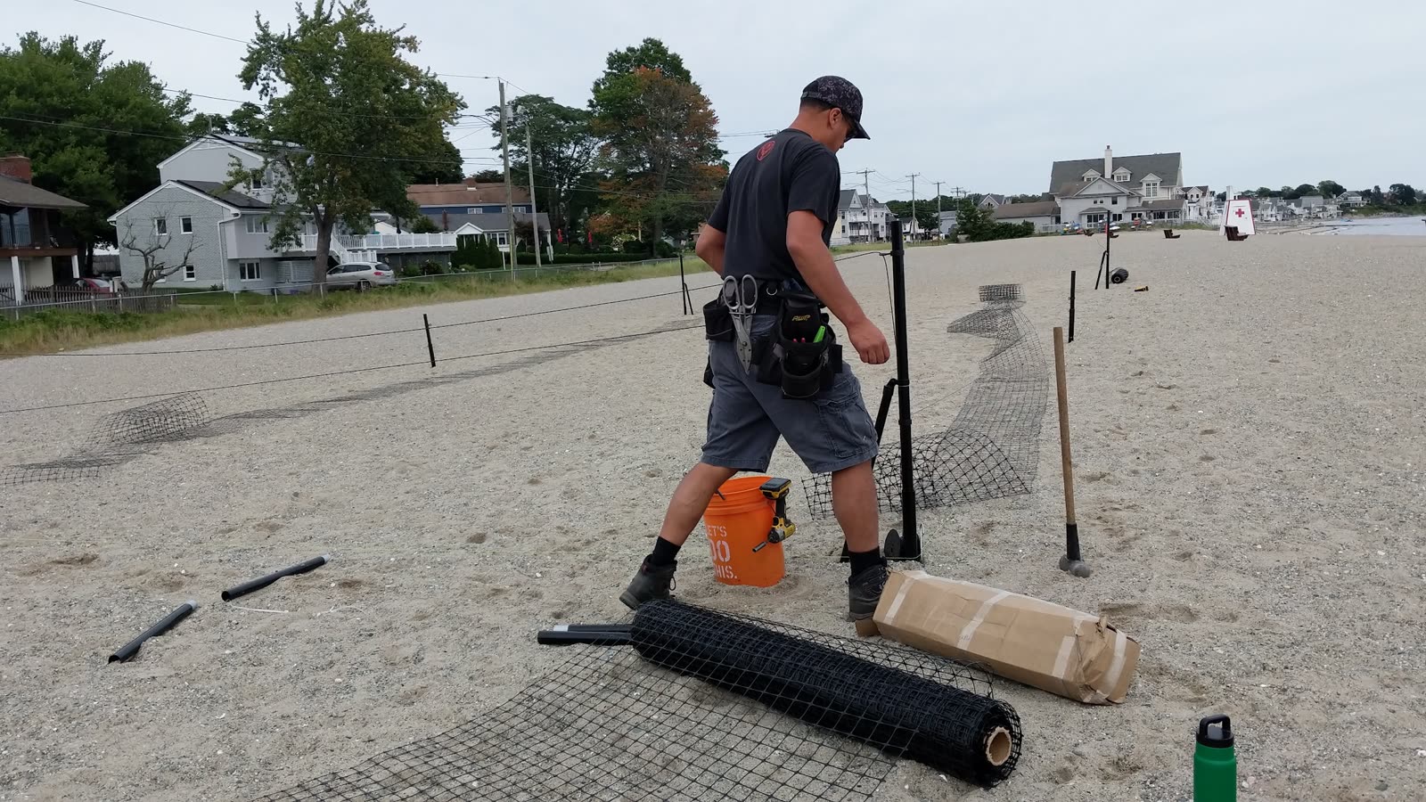 A man carrying a post across a beach with rolled black netting and a bucket of hardware on the sand
