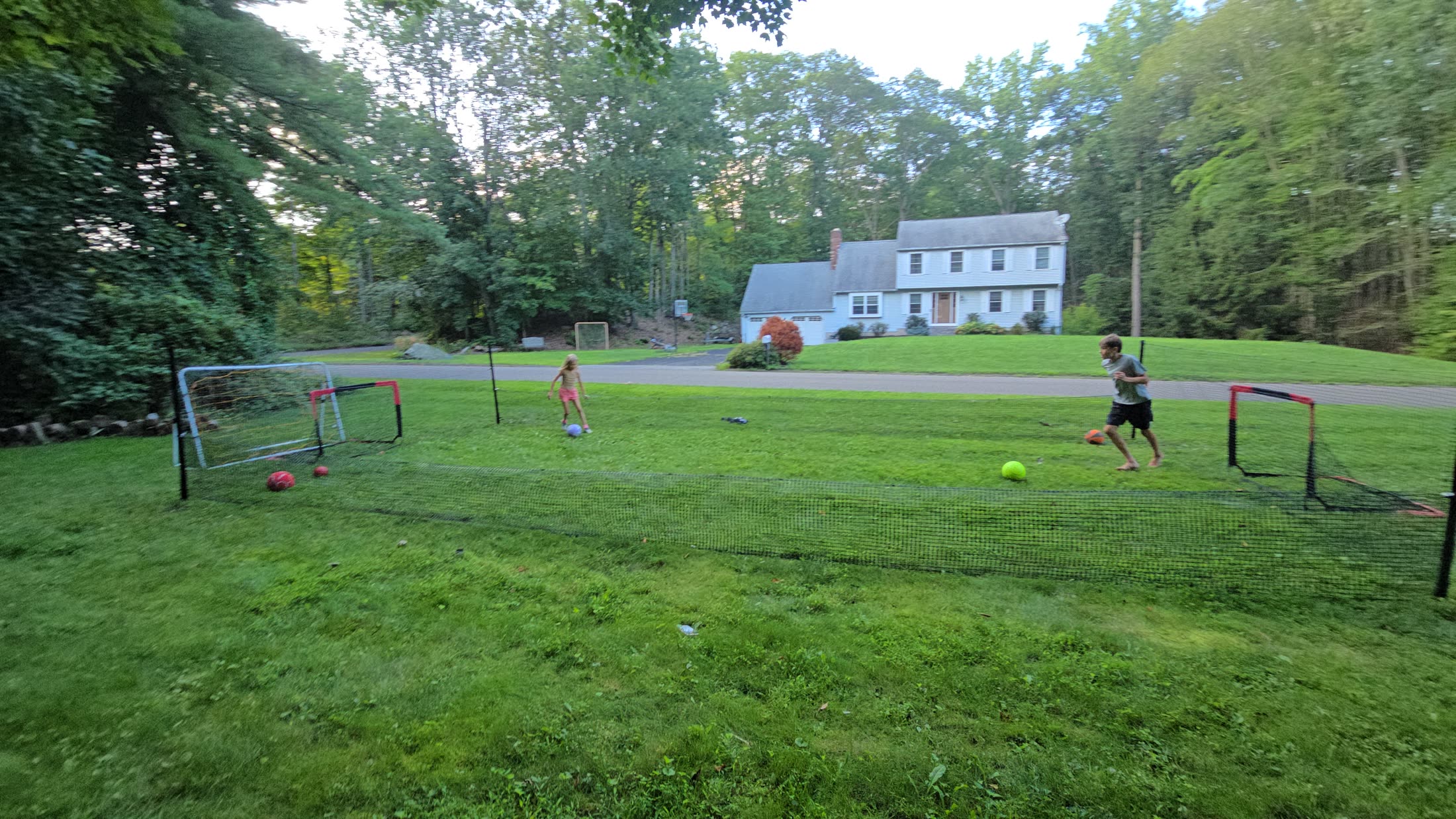Wide view of the finished enclosed field with goals at both ends and two children playing, a house beyond the road