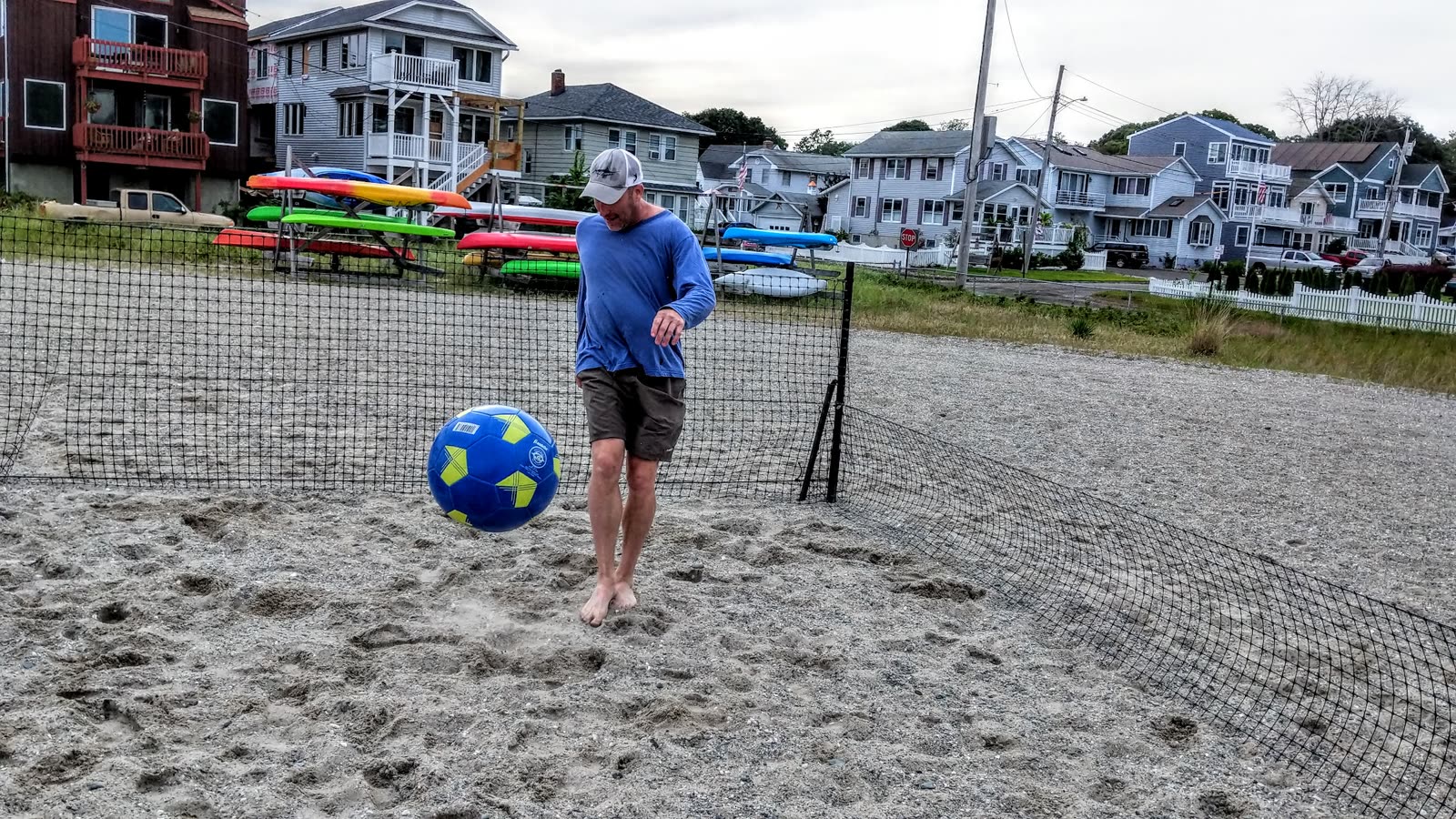A man standing with a blue-and-yellow ball inside the netted beach field, kayaks and houses behind
